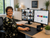 A woman with short curly hair and glasses sits at a desk in a bright office, smiling at her dual computer monitors. One screen shows a video conference with multiple participants, and the other displays a dashboard.