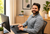 A smiling man with a beard sits at a desk using a computer keyboard and mouse in a bright home office setting.